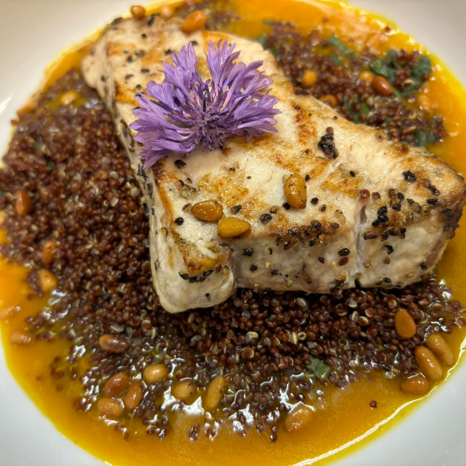 Grilled fish with a purple flower garnish, served over quinoa, pine nuts, and a yellow sauce.
